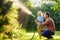 Funny little boy with his father watering plants and playing with garden hose in sunny backyard. Preschooler child having fun with