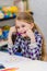 Funny little girl with blond hair sitting at table and holding purple pencil in her mouth