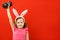 Funny little girl in easter bunny ears raised up a dumbbell. A child on a red background color