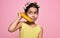 A funny little girl holds a banana, like a phone, isolated on pink background. A happy kid in a yellow dress pretends to talk on a