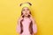 Funny little girl with ponytails wearing pink shirt and beanie hat standing isolated over yellow background pointing at her pout