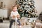 Funny little girl preschooler with blond hair in a white sweater rides a rocking horse. Christmas tree in the background