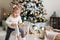 Funny little girl preschooler with blond hair in a white sweater rides a rocking horse and smiles. Christmas tree in the