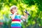 Funny little kid boy shooting wooden slingshot against green tree background. Child having fun in summer