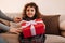 Funny little kid holding birthday present. Curly preteen girl with gift sitting on sofa