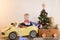 Funny little smiling kids driving toy car with Christmas tree. Happy child in colour fashion clothes bringing hewed xmas tree from