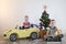 Funny little smiling kids driving toy car with Christmas tree. Happy child in colour fashion clothes bringing hewed xmas tree from