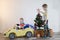 Funny little smiling kids driving toy car with Christmas tree. Happy child in colour fashion clothes bringing hewed xmas tree from