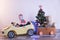 Funny little smiling kids driving toy car with Christmas tree. Happy child in colour fashion clothes bringing hewed xmas tree from