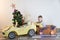 Funny little smiling kids driving toy car with Christmas tree. Happy child in colour fashion clothes bringing hewed xmas tree from