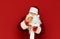 Funny man in Santa Claus costume stands on red background with burger in hand, looks at fast food with surprised face. Shocked