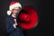 Funny mischievous child boy in Santa hat speaking through a loudspeaker on blackboard background. Kid with red megaphone