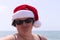 Funny portrait of a overweight woman in a santa hat and sunglasses against the background of the sea.