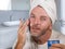 Funny portrait of young happy and attractive camp gay man in bathroom applying moisturizer facial cream with head wrapped in towel