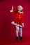 Funny schoolgirl in Xmas costume with Santa`s hat jumping on red colored background with plenty of space. Kid points up.