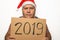 Funny senior serious man in red Santa Claus Christmas hat holds cardboard with 2019 numbers in hands and angrily looks at camera