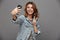 Funny teen girl in jeans jacket showing peace gesture while taking selfie on smartphone