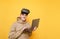 Funny teenager in casual clothes stands on a yellow background with a laptop in his hands with VR glasses on his head, looks into