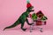 funny toy dinosaur wearing tiny knitted hat and driving shopping trolley full of present boxes on a pink background ,