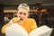 Funny young man in a cap bites a sandwich and reads a book with a focused look. Guy in a yellow sweater learns at a cafe. Cheerful