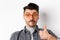 Funny young man in glasses show thumb up, biting lip and look indecisive, standing on white background