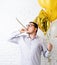 Funny young man wearing birthday hat and holding balloons blowing noisemaker celebrating birthday