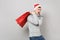 Funny young Santa man in Christmas hat hold red package bag with gifts, purchases after shopping on grey