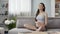 Future mother sitting on sofa meditating, touching stomach, healthy pregnancy