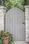 Garden gate in rock wall with pot of geraniums to one side and trees and vines visible over the top