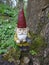 Garden gnome with an ax in his hand stands in the woods on a tree stump between green plants.