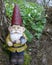 Garden gnome with an ax in his hand stands in the woods on a tree stump between green plants.