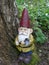 Garden gnome with an ax in his hand stands in the woods on a tree stump between green plants.