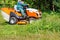 A gardener mows tall green grass with a tractor lawn mower