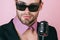 Gay man in glasses sing in microphone. Sexy dj young man on pink background. Music, look and retro style. Freak singer