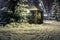 Gazebo, wooden house and Christmas trees in the snow at night. A trodden door leads to the gazebo. Lanterns and garlands