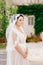 A gentle bride in a lace veil stands near a white house entwined with a vine in the old town of Perast