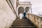 Gentle groom holding hand of his pretty bride when descending by antique palace stone stairs. Low angle shot