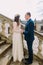 Gentle groom holding hands with his pretty bride while both stand on antique stone stairs