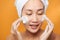 Gentle half-naked woman wrapped in towel washing her face with foaming cleanser isolated over orange background