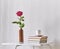 Gentle pink rose in clay bottle, cup of coffee, books on a white coffee table against a light wall.
