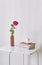 Gentle pink rose in clay bottle, cup of coffee, books on a white coffee table against a light wall.