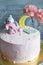 Gently pink children`s cake with a little gray bear hugging a pink blanket and a yellow crescent on a gray blurry background with