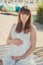 Genuine cute pregnant lady woman in white airy dress sitting sand beach wooden palette bridge holding tummy abdomen. Attractive be
