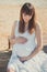 Genuine cute pregnant lady woman in white airy dress sitting sand beach wooden palette bridge holding tummy abdomen. Attractive be