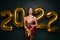 Georgeous brunette lady in beautiful dress holding gift box on black studio background with golden 2022 balloons