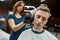 Getting new trendy haircut. Young barber girl or female hairdresser working with hair clipper, serving young guy sitting