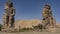 Giant sculptures of the colossi of Memnon against a blue sky and a sand dune.