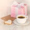 Gift box wrapped in pink dotted paper, heart shaped love cookie, a cup of coffee and an empty kraft card over a white wood