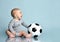 Ginger child in gray bodysuit, barefoot. He holding soccer ball, sitting on floor against blue background. Close up, copy space