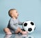 Ginger child in gray bodysuit, barefoot. He holding soccer ball, sitting on floor against blue background. Close up, copy space